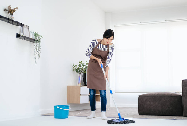 Pretty young lady in apron wiping floor with mop, doing house cleanup living room, free space. Positive housewife tidying her apartment, doing household chores. Professional sanitary service concept. - Valokuva, kuva