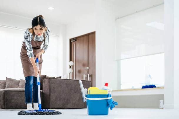 Pretty young lady in apron wiping floor with mop, doing house cleanup living room, free space. Positive housewife tidying her apartment, doing household chores. Professional sanitary service concept. - Foto, Bild