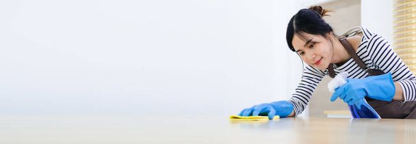 Woman in protective gloves is smiling and wiping dust using a spray and a duster while cleaning her house, close-up - Фото, изображение