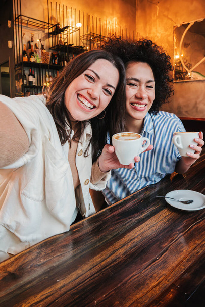 Retrato vertical de duas jovens mulheres adultas se divertindo tirando uma selfie no café sentado à mesa com xícaras. Par de fêmeas alegres sorrindo no restaurante. Duas raparigas a divertir-se juntas. Alto - Foto, Imagem