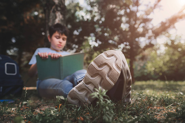 Selective focus on the sole of the sneakers of a handsome smart teenage schoolboy, sitting near his backpack under a tree in a pine park, reading a book at sunset. Education. Childhood. Adolescence - Zdjęcie, obraz
