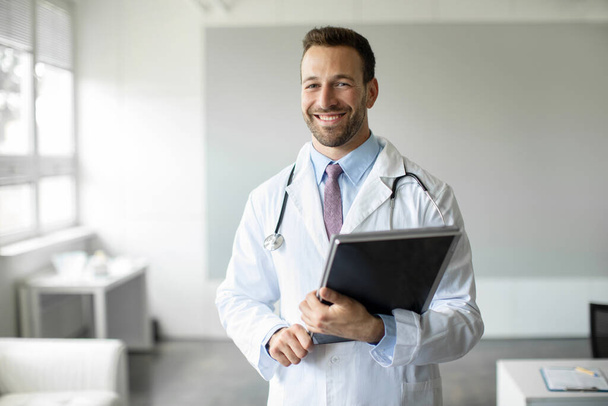 Πορτρέτο του χαρούμενου ανθρώπου γιατρός κρατώντας πρόχειρο χαμογελώντας στην κάμερα, αρσενικό γιατρό GP που εργάζονται στην κλινική, φορώντας λευκή ιατρική στολή, αντίγραφο χώρο - Φωτογραφία, εικόνα
