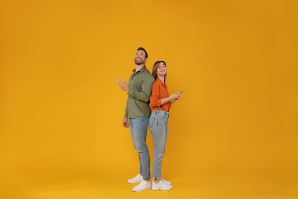 Full body length shot of two friends man and woman standing back to back, holding cellphones and looking up at free space, browsing internet isolated on yellow background - Photo, Image