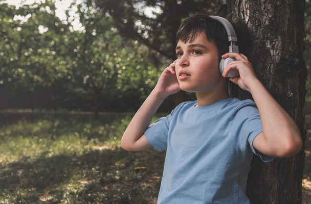 Retrato dentado de um adolescente de cabelos escuros caucasiano adorável, estudante de camiseta casual azul, ouvindo música em fones de ouvido sem fio, relaxando após a aula, sentado sob uma árvore no parque - Foto, Imagem