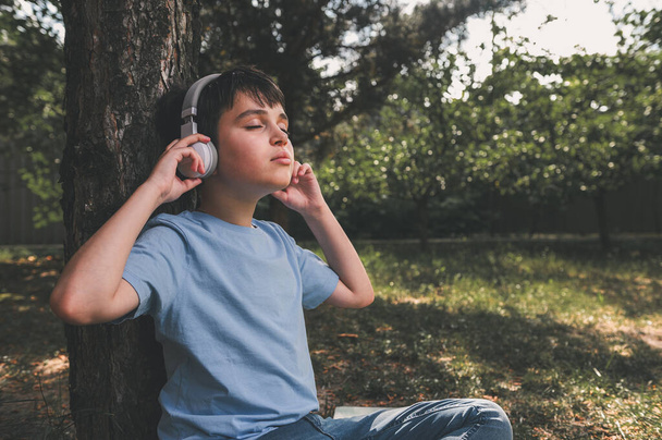 Retrato dentado de um adolescente caucasiano adorável com fones de ouvido sem fio, desfrutando de nova trilha sonora enquanto descansa no parque ao pôr do sol, sentado debaixo da árvore. Crianças entretenimento estilo de vida Hobby - Foto, Imagem