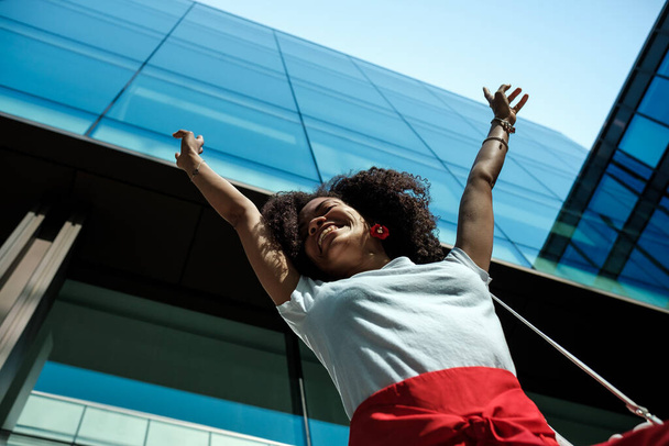 Heureuse femme noire rit les bras en l'air devant un immeuble en verre cosmopolite. Bonheur et rire concept. - Photo, image