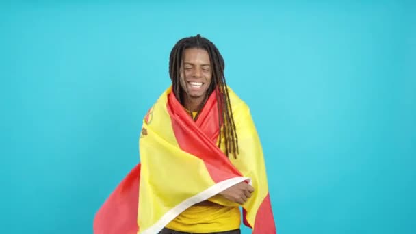 Stüdyoda mavi arka planda İspanyol bayrağıyla sarılan rastalı mutlu Latin adam. - Video, Çekim