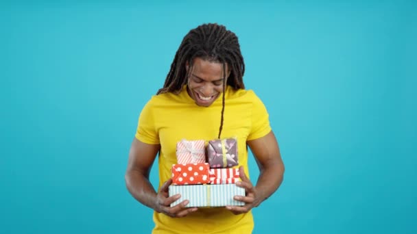Ευτυχισμένος Λατίνος άνθρωπος με dreadlocks κρατώντας πολλά πολύχρωμα δώρα στο στούντιο με μπλε φόντο - Πλάνα, βίντεο