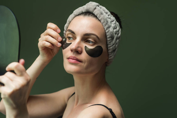 Attractive European woman puts patches under her eyes, looking in the mirror, on a green background. Mature skin care, hydrogel patches under the eyes. - Photo, Image