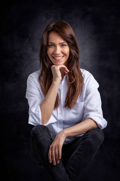 Attractive middle aged woman with toothy smile wearing white shirt and black jeans while sitting against at isolated dark background. Copy space. Studio shot. - Fotó, kép