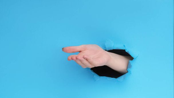 Give gesture. Pay money. Female hand open palm inside breakthrough hole on blue ripped paper wall advertising background with empty space. - Zdjęcie, obraz
