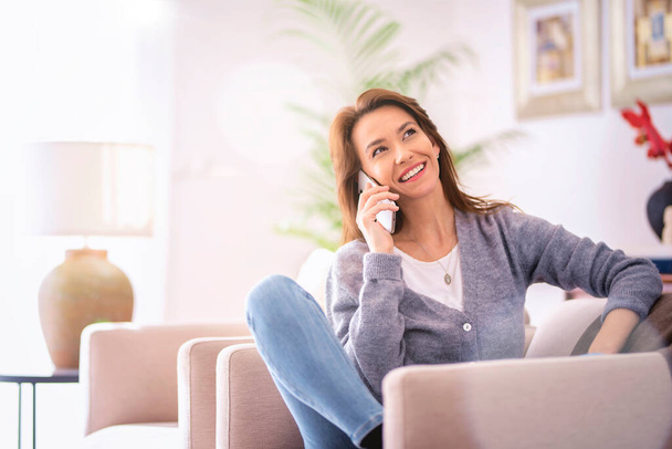 Brunette haired woman relaxing in an armchair at home and having a call. Confident female wearing casual clothes and cheerful smiling.  - Foto, Imagen