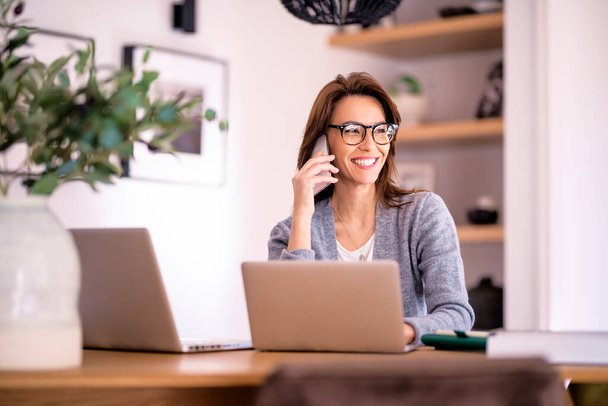 Attractive woman wearing eyewear and casual clothes while working from home. Business woman using laptops and making a call. Home office. - Fotó, kép