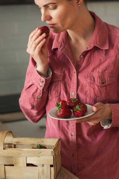 Una mujer recoge fresas de una cesta de pie en la cocina. Fresas en manos de una mujer Una mujer toma fresas de una canasta - Foto, imagen