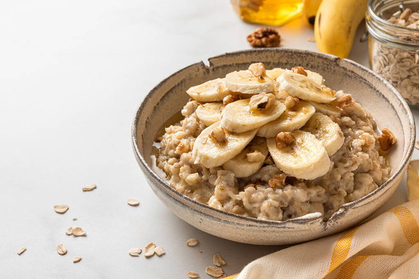 Healthy breakfast. Bowl of oatmeal porridge with banana, walnuts and honey on white table. Close up. Healthy diet food - Photo, Image
