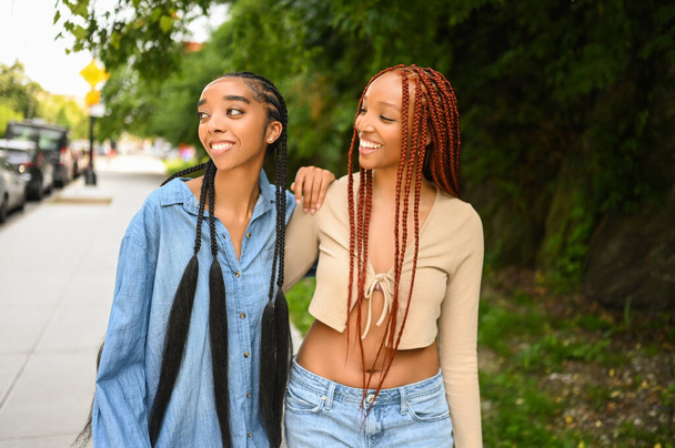ストリートスタイルの肖像画美しい若いアフリカ系アメリカ人女性の黒のカップル。毛、完璧な白い歯の笑顔、夏の晴れた日に屋外を歩いて抱擁姉妹の友人。緑の公園と都市 - 写真・画像