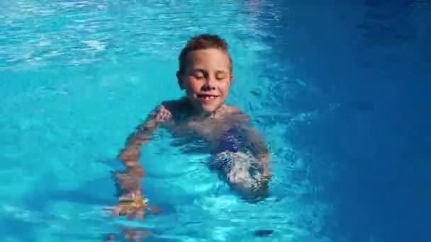 8-9-vuotias poika ui uima-altaassa sinisen veden kanssa. Kesälomat, lomat. Lasten terveyden parantaminen - Materiaali, video