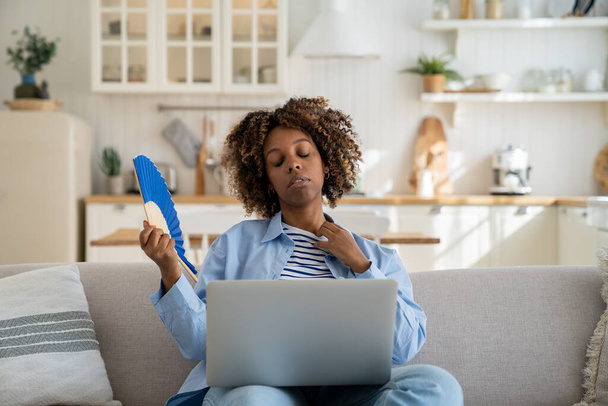 Sweaty African American millennial girl suffer from heat while working remotely at home, sitting on sofa with laptop on knees and waving hand fan, trying to cool off at home during extreme hot weather - Photo, Image