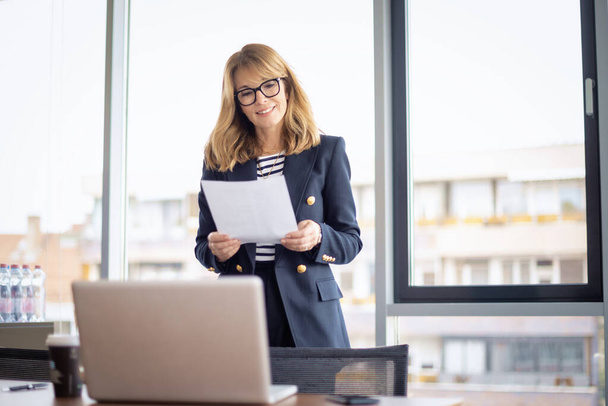 Shot of a confident mature businesswoman standing in a modern office and holding document in her hand. Professional female wearing eyeglasses and blazer. - Valokuva, kuva