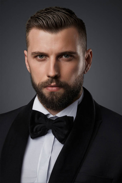 Nahaufnahme Porträt eines stilvollen und attraktiven Mannes mit gepflegtem Bart, bekleidet mit einem hochwertigen schwarzen Anzug und Fliege, der selbstbewusst vor grauem Hintergrund posiert - Foto, Bild