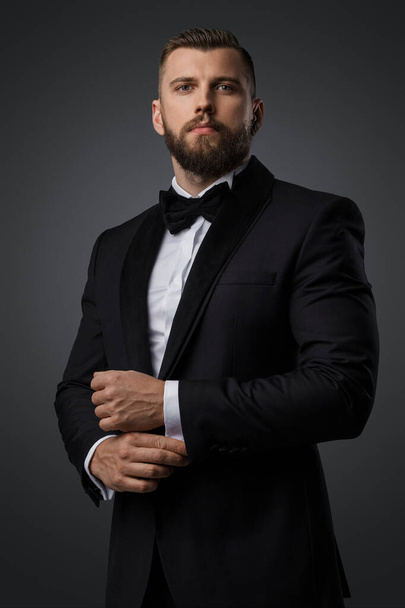 Confident and stylish handsome man with a manicured beard wears a luxurious black suit and bowtie on a grey background - Valokuva, kuva