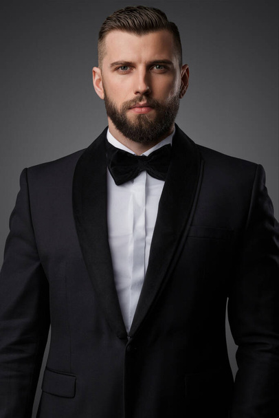 Confident and stylish handsome man with a manicured beard wears a luxurious black suit and bowtie on a grey background - Фото, изображение