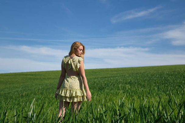 Junge, schöne, blonde Frau in gelbem Kleid, die durch ein grünes Weizenfeld geht, allein, ruhig und friedlich. Konzept Schönheit, Entspannung, Ruhe, Ruhe, Felder, Wiesen. - Foto, Bild