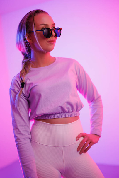 Giovane donna sicura di sé e cool che indossa occhiali da sole in piedi con mano sul fianco contro sfondo viola illuminato - Foto, immagini