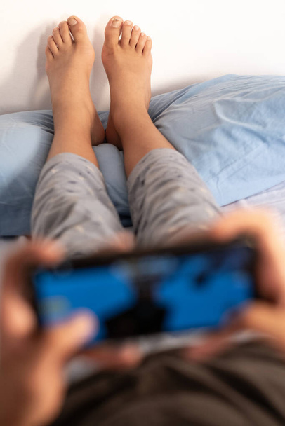 Um adolescente de pijama no meio da manhã deitado na cama está jogando videogames com os pés apoiados na almofada. Concentre-se no espaço de fundo e cópia. - Foto, Imagem