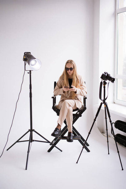 Una productora de video, una asistente rubia con cámara y smartphone, trabajando en el teléfono en el estudio. El uso de un traje de pantalón desnudo formal sobre un fondo blanco aislado.  - Foto, imagen