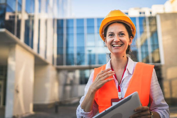 architekt kobieta inżynier budownictwa stoją na zewnątrz nosić kask ochronny i zachód przed nowoczesną ścianą budynku - Zdjęcie, obraz