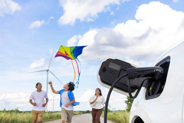 Concept of progressive happy family holding windmill toy and relax at wind farm with electric vehicle. Electric vehicle driven by clean renewable energy from wind turbine generator to charger station. - Photo, image
