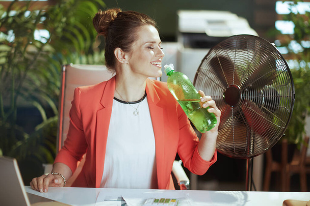Sustainable workplace. happy modern business woman at work in red jacket with bottle of water and electric fan. - Φωτογραφία, εικόνα