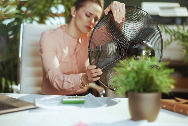 Sustainable workplace. modern middle aged bookkeeper woman at work with electric fan suffering from summer heat. - Photo, Image