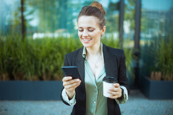 happy modern business woman near office building in black jacket with cup of coffee using smartphone. - Foto, immagini