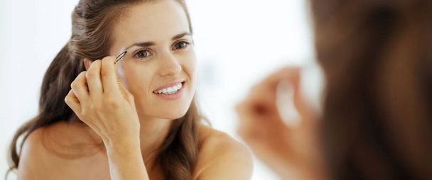 Happy young woman shaping eyebrows in bathroom - Foto, Imagen