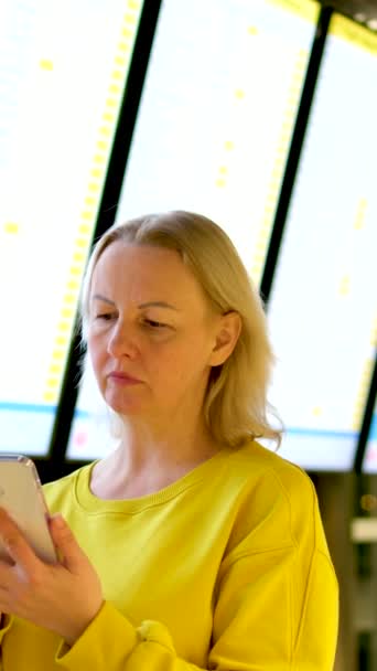 επίλυση προβλημάτων στο αεροδρόμιο μια γυναίκα στα κίτρινα στέκεται μπροστά από την τηλεόραση και μιλάει στο τηλέφωνο σοβαρό πρόσωπο που περιμένει - Πλάνα, βίντεο