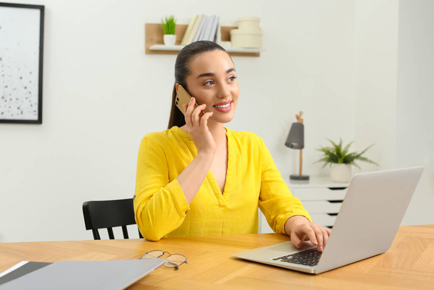 Home workplace. Happy woman talking on smartphone near laptop at wooden desk in room - Fotoğraf, Görsel