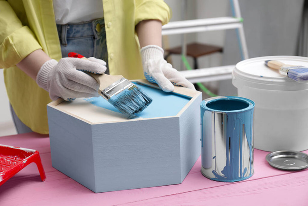 Woman painting honeycomb shaped shelf with brush at pink wooden table indoors, closeup - 写真・画像