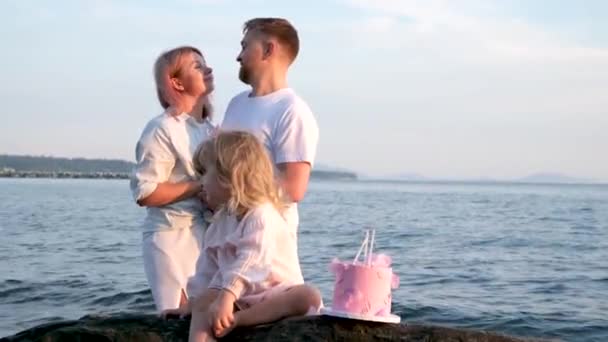 Любіть першу дитину в сімейній дівчині-блондинці з тортом 3 роки любові між батьками, які обіймаються, дивляться один на одного на камені біля бороди океанів. Світло - білий одяг - справжня прекрасна сім "я. - Кадри, відео