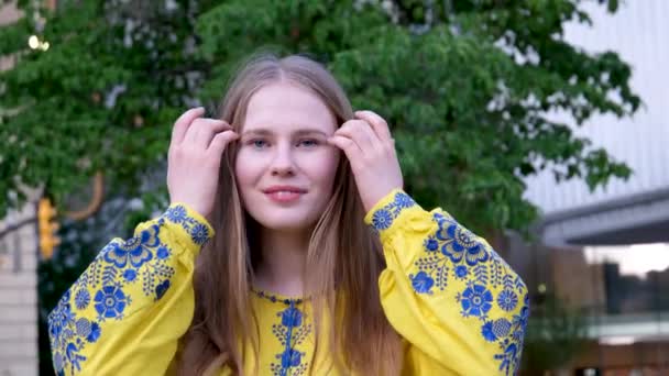 young Ukrainian woman embroidered shirt looks into smiles straightens. Hair blond hair blurred background close-up face earrings in the form of flag of Ukraine Victory fun joy strength Freedom Will - Footage, Video