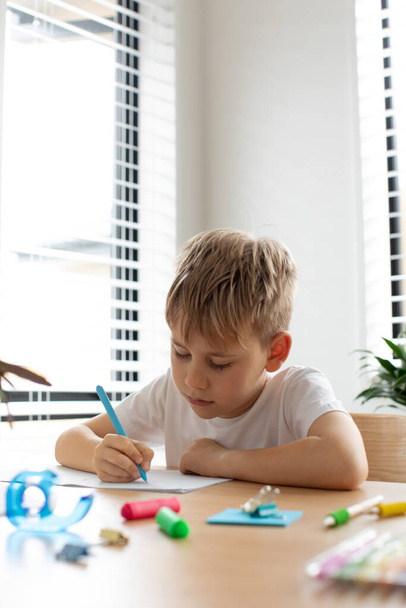 Cute boy doing school homework at home. Distance education. Online education. The boy is writing intently in a notebook. - Zdjęcie, obraz