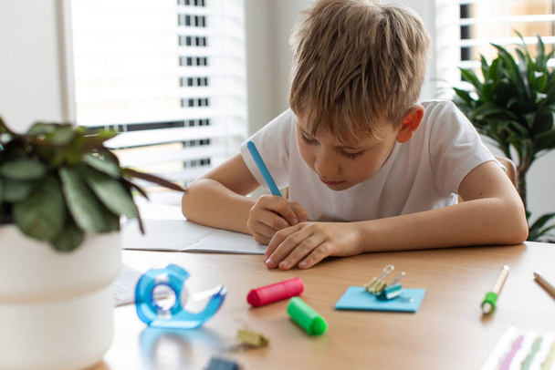 Cute boy doing school homework at home. Distance education. Online education. The boy is writing intently in a notebook. - Valokuva, kuva
