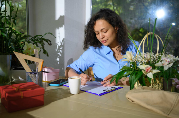 Mooie vrouw, bloemist ondernemer, ondernemer, event manager schrijven op klembord, werken met bestellingen in een gezellige kleine bloemenwinkel. Bloemenarrangementen en geschenken voor feestelijke evenementen - Foto, afbeelding