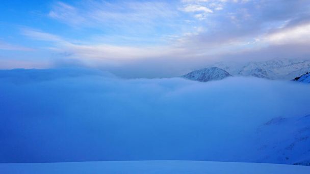 Een oceaan van wolken in de besneeuwde bergen bij dageraad. Witte wolken zijn als een tapijt in een kloof. Golven stijgen en dalen af van de bergen. Zonsopkomst van de gele zon. Heldere lucht, bergtoppen. Almaty - Foto, afbeelding