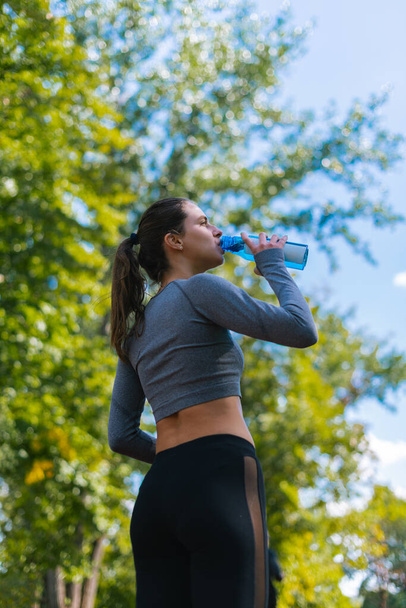 Молодая, красивая девушка стоит и пьет воду после тренировки в парке - Фото, изображение