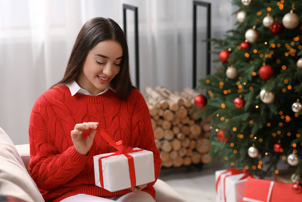 Smiling woman opening gift near Christmas tree indoors - 写真・画像