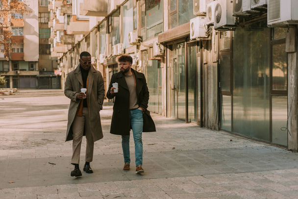 A front view photo of two stylish multiracial businesspeople going to work and having a conversation, on a beautiful sunny morning. They are holding cups of coffee - Фото, изображение