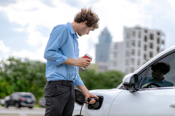 Progressive eco-friendly concept of parking EV car at public electric-powered charging station in city with blur background of businessman leaning on recharging-electric vehicle with coffee. - Foto, afbeelding