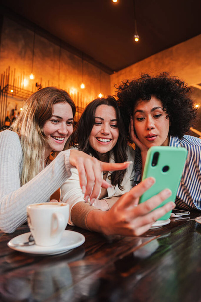 Vertical portrait of a group of young women having fun sharing media with an cellphone. Females looking the smartphone on a coffee shop. Girls shopping online on a marketplace using a mobile phone - 写真・画像